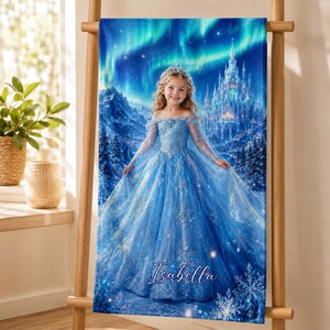 Op de afbeelding: Een blauwe strandlaken met een jong meisje in een blauwe jurk, een tiara en een kasteel in een winterlandschap. De achtergrond toont het noorderlicht. De naam "Isabella" staat onderaan in cursief.