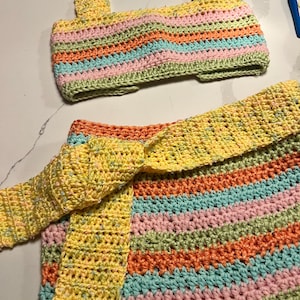 May include: A two-piece crochet set consisting of a sleeveless crop top and a skirt with a tie-waist belt. The garment features horizontal stripes in pastel pink, light blue, and orange, accented by a speckled yellow yarn throughout the design.