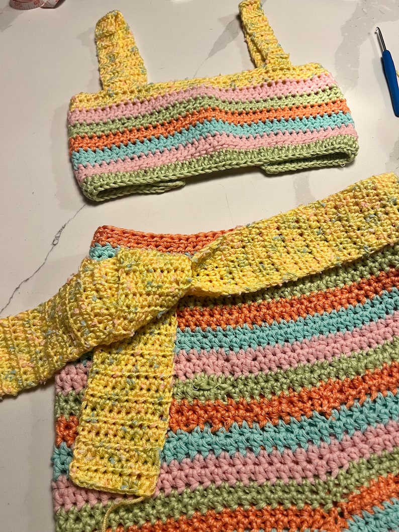 May include: A two-piece crochet set consisting of a sleeveless crop top and a skirt with a tie-waist belt. The garment features horizontal stripes in pastel pink, light blue, and orange, accented by a speckled yellow yarn throughout the design.