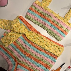 May include: A two-piece crochet set consisting of a sleeveless crop top and high-waisted shorts. Both pieces feature horizontal stripes in pastel yellow, orange, pink, and light blue. The shorts include a matching tie-waist belt.