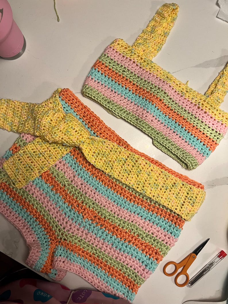 May include: A two-piece crochet set consisting of a sleeveless crop top and high-waisted shorts. Both pieces feature horizontal stripes in pastel yellow, orange, pink, and light blue. The shorts include a matching tie-waist belt.