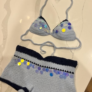 May include: A two-piece crochet set consisting of a light blue bikini top and matching shorts. Both pieces feature dark navy blue trim and are embellished with iridescent, circular sequins that reflect various colors across the light blue yarn.