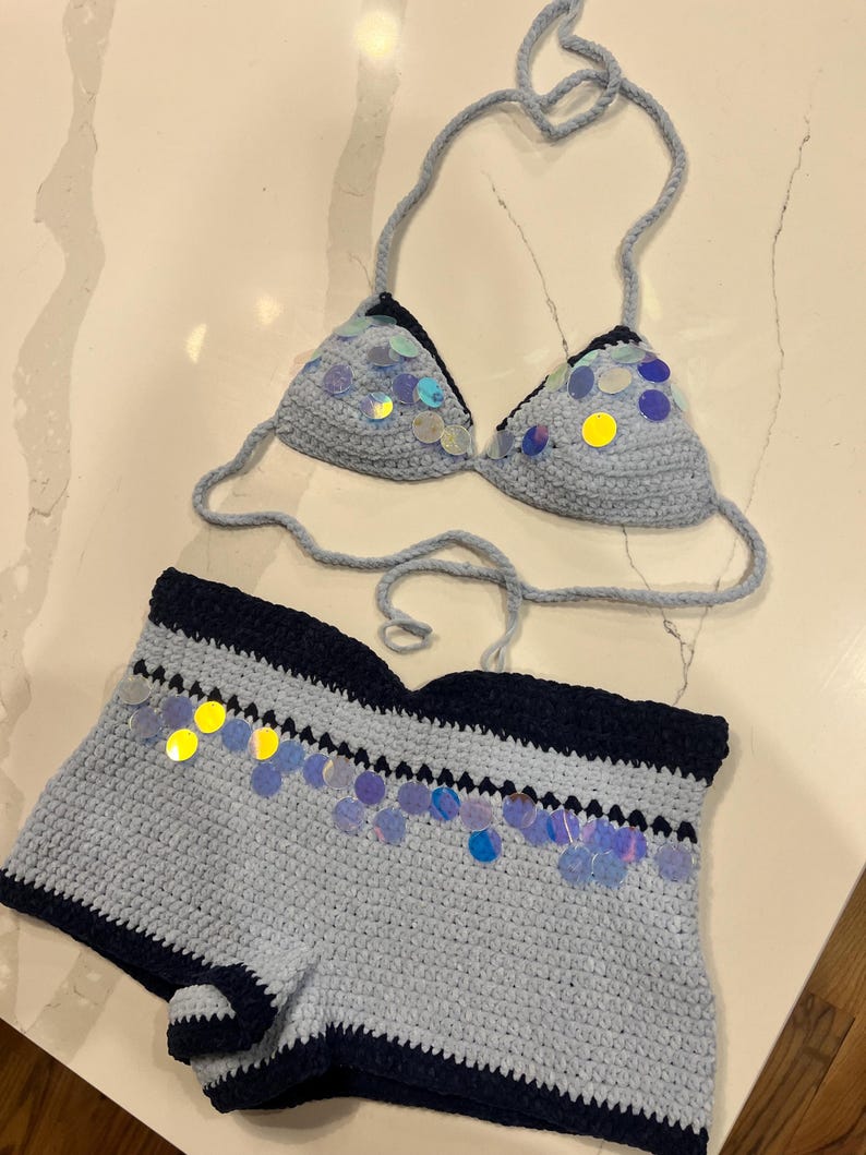 May include: A two-piece crochet set consisting of a light blue bikini top and matching shorts. Both pieces feature dark navy blue trim and are embellished with iridescent, circular sequins that reflect various colors across the light blue yarn.