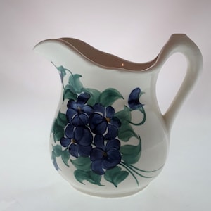 May include: White ceramic pitcher with a curved handle and spout. The pitcher is decorated with a cluster of blue flowers and green leaves. The rim has a light brown trim.