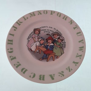 May include: A white ceramic plate with a green border and the alphabet printed around the edge. The center of the plate features a colorful illustration of children playing music. The text on the plate reads "Tom he was a piper's son... he learned when he was young to play."