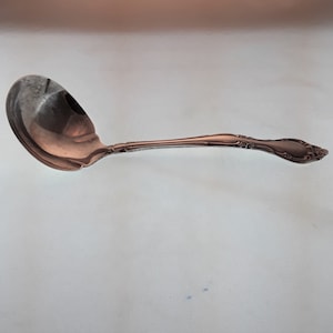 May include: A silver ladle with an ornate handle. The ladle is designed for serving liquids, such as soup or gravy.