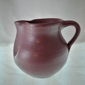 May include: A deep burgundy ceramic pitcher with a rounded body and a slightly flared lip. The pitcher has a simple, elegant design and a smooth, glossy finish.