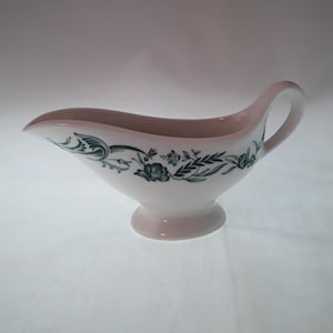 May include: White ceramic gravy boat with a green floral design. The boat has a wide, rounded base and a spout on one side.