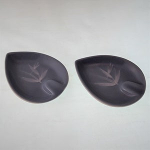 May include: Two small, gray ceramic ashtrays with a leaf design. Each ashtray has a small indentation for cigarettes.