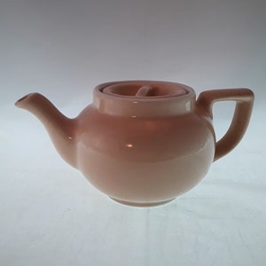May include: A pale pink ceramic teapot with a lid and a handle. The teapot has a rounded body and a spout.