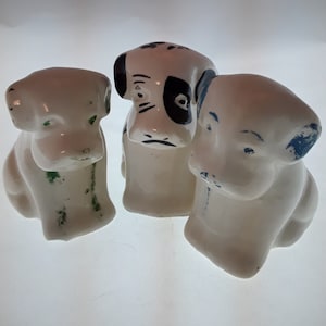 May include: Three ceramic dog figurines, each with a different design. One is white with black spots and blue accents, another is white with green accents, and the third is white with blue accents.