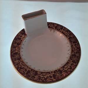 May include: A white ceramic dish with a gold rim and a floral design in shades of brown and gold. The dish has a raised rectangular section on one side.