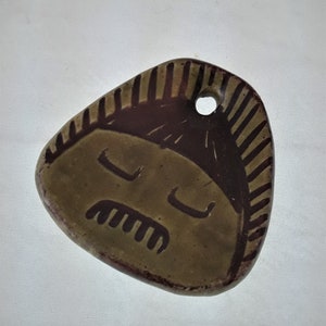 May include: A brown ceramic ornament with a stylized face and a hole for hanging. The face has a mustache and closed eyes.
