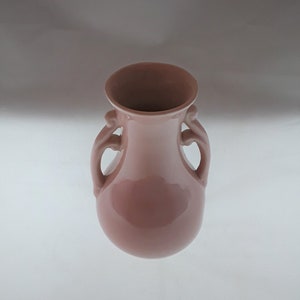 May include: A pink ceramic vase with two curved handles on either side. The vase has a rounded body and a flared opening. The color is a muted, dusty rose, and the surface has a glossy finish. The vase is likely for decorative purposes.