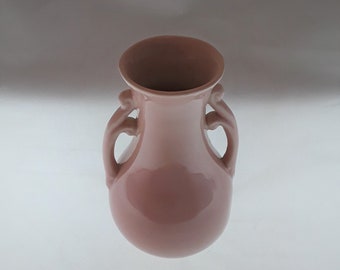 May include: A pink ceramic vase with two curved handles on either side. The vase has a rounded body and a flared opening. The color is a muted, dusty rose, and the surface has a glossy finish. The vase is likely for decorative purposes.