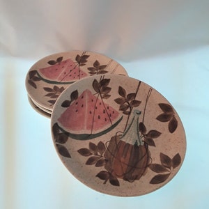 May include: Set of four vintage ceramic dinner plates with a brown, green, and pink watermelon and bottle design. The plates have a speckled beige background.
