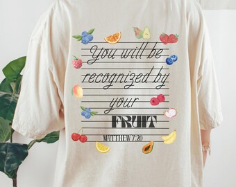 May include: Oatmeal-coloured t-shirt featuring a fruit and text design on the back. The text says "You will be recognised by your FRUIT" with "Matthew 7:20" underneath. Watercolour illustrations of various fruits surround the text.