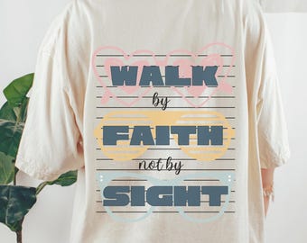 May include: A cream-coloured t-shirt featuring the phrase "WALK by FAITH not by SIGHT". The text is in a layered design with different colours and styles, framed by decorative elements. The t-shirt is a neutral colour.