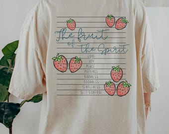 May include: A cream-coloured t-shirt featuring the phrase "The fruit of the Spirit" in teal script, with strawberry illustrations. The shirt lists virtues such as love, joy, and peace. The design is on the back.