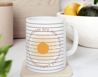 May include: White ceramic mug with a wraparound design featuring a stylized sunburst in orange and yellow, encircled by the text "His mercies are new every morning, Lamentations 3:22". The mug has a simple handle and horizontal black lines.