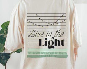 May include: Cream-coloured t-shirt with a graphic on the back. The design features the words "Live in the Light" in a stylized font, with a string of lights above and a quote below. The shirt has a relaxed fit.
