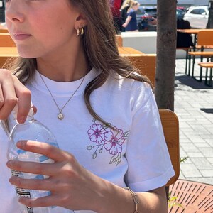 May include: A white t-shirt with a floral design in purple and green. The person is wearing gold hoop earrings, a gold heart pendant necklace, and a gold chain bracelet. A clear water bottle is held in the hand.