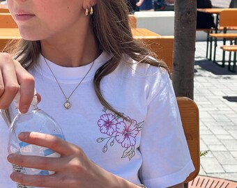 May include: A white t-shirt with a floral design in purple and green. The person is wearing gold hoop earrings, a gold heart pendant necklace, and a gold chain bracelet. A clear water bottle is held in the hand.