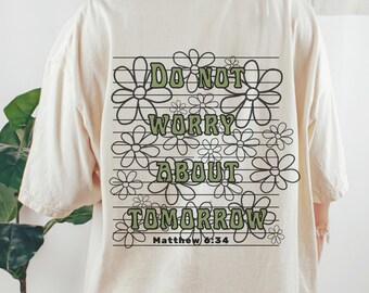 May include: A cream-coloured t-shirt featuring a floral design and the green text "DO NOT WORRY ABOUT TOMORROW". The text is surrounded by daisy outlines, with "Matthew 6:34" below.