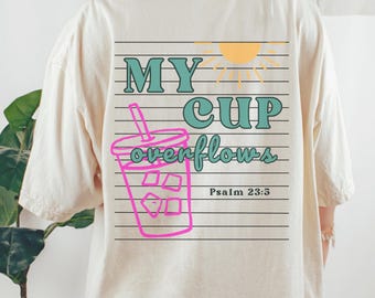 May include: Cream-coloured t-shirt with a graphic on the back. The graphic features the text "MY CUP overflows" in teal and a pink drink illustration. A yellow sun and the Bible verse Psalm 23:5 are also included.