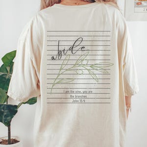 May include: Cream-colored t-shirt with a graphic on the back. The graphic features the word "abide" in cursive, olive branch illustration, and the text "I am the vine, you are the branches John 15:4" in a rectangular design.