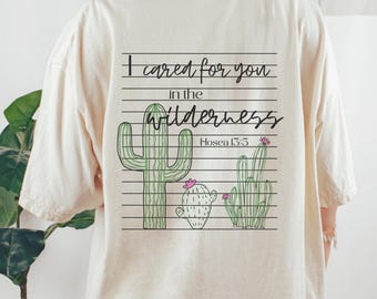 May include: A cream-coloured t-shirt with a graphic of three green cacti and pink flowers. The text "I cared for you in the wilderness Hosea 13:5" is printed above the cacti in black. The shirt has a loose fit.