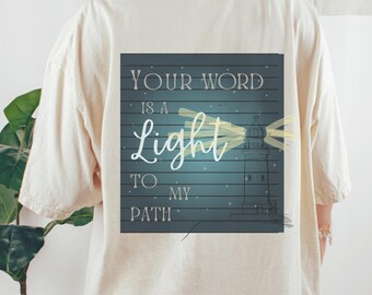 May include: A cream-coloured t-shirt with a square graphic on the back. The graphic shows a lighthouse emitting light, with the words "YOUR WORD IS A LIGHT TO MY PATH" in white script and block letters.