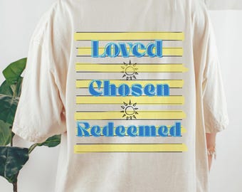 May include: A cream-coloured t-shirt featuring the words "Loved, Chosen, Redeemed" in blue, set against yellow horizontal stripes. The design includes sun graphics. The shirt has a relaxed, oversized fit.