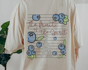 May include: A cream-coloured t-shirt with a graphic on the back. The design features blueberries and the text "The fruit of the Spirit." The words "Love, Joy, Peace, Patience, Kindness, Goodness, Gentleness, Self Control" are also included.