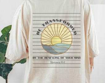 May include: Cream-coloured t-shirt with a graphic on the back. The graphic features a sunburst design with the words "BE TRANSFORMED BY THE RENEWING OF YOUR MIND" and the text "Romans 12:2".