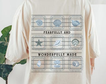 May include: A cream-coloured t-shirt with a grid of light blue seashell illustrations, each in a decorative frame. The text "FEARFULLY AND WONDERFULLY MADE" is printed below. The shirt has a loose fit.