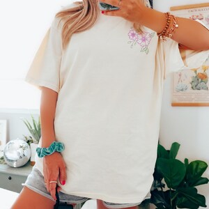 May include: Cream-coloured t-shirt featuring a floral design in purple and green. The short-sleeved shirt is made of a soft material. The floral design is on the upper left side of the shirt. The shirt is a neutral colour.