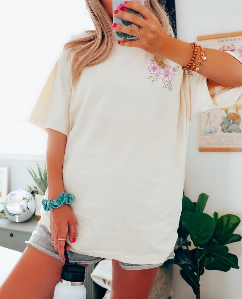 May include: Cream-coloured t-shirt featuring a floral design in purple and green. The short-sleeved shirt is made of a soft material. The floral design is on the upper left side of the shirt. The shirt is a neutral colour.