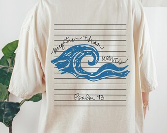 May include: A cream-coloured t-shirt featuring a blue wave design with the text "mightier than waves" and "Psalm 93". The graphic is set against a lined backdrop.