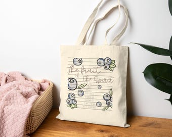 May include: A cream-coloured tote bag featuring the words "The Fruit of the Spirit" and a blueberry illustration. The bag lists virtues such as love, joy, peace, and kindness. The design is simple, with a natural aesthetic.