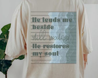 May include: A cream-coloured t-shirt with a rectangular graphic on the back. The graphic features the text "He leads me beside still waters He restores my soul" in a grey font, with a blue water design.