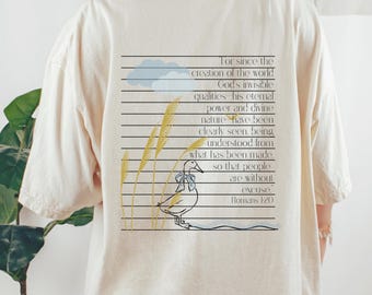 May include: A cream-coloured t-shirt with a graphic design on the back. The design features a duck with a blue bow, wheat stalks, and text from Romans 1:20. The text is arranged in horizontal lines.