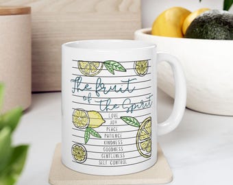 May include: White ceramic mug featuring the words "The Fruit of the Spirit" in a script font, accompanied by a list of virtues. The design incorporates illustrations of lemons and leaves. The mug rests on a beige coaster.