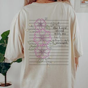 May include: A cream-coloured t-shirt with a floral design and text on the back. The design features pink flowers with green leaves and the words "But the Lord stood with me and gave me strength" from 2 Timothy 4:17.
