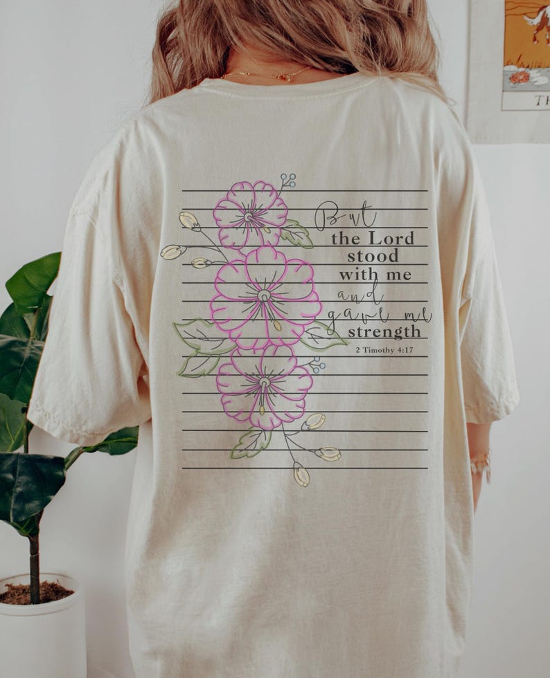 May include: A cream-coloured t-shirt with a floral design and text on the back. The design features pink flowers with green leaves and the words "But the Lord stood with me and gave me strength" from 2 Timothy 4:17.