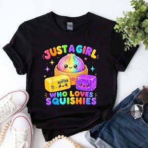 Puede incluir: Una camiseta negra con un gráfico colorido de una bola de masa arcoíris en una vaporera de bambú, una barra de mantequilla amarilla con la etiqueta 4oz y un bloque cuadrado morado. El texto dice JUST A GIRL WHO LOVES SQUISHIES en letras brillantes.