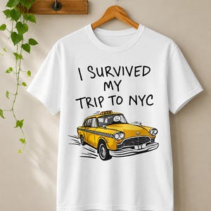 May include: A white short-sleeve t-shirt featuring the black text I SURVIVED MY TRIP TO NYC above a graphic illustration of a yellow taxi cab with a stick figure driver. The taxi has a checkered side stripe and a roof sign reading TAXI.