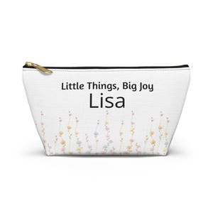 May include: White zippered pouch with the text "Little Things, Big Joy Lisa". The pouch features a floral design with pastel-colored flowers at the bottom and a gold-colored zipper.