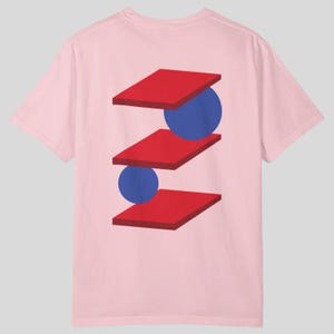 May include: A light pink t-shirt featuring a geometric design on the back. The design consists of three stacked red rectangles with blue circles between them. The shirt has short sleeves and a crew neck.