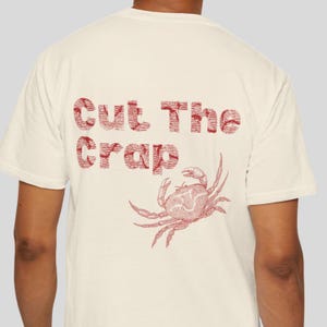 以下が含まれることがあります： 赤色のカニのイラストと「Cut The Crap」の文字が入ったクリーム色のTシャツ。カニの細密なイラストと、かすれた赤い文字が特徴で、ヴィンテージ感を演出しています。